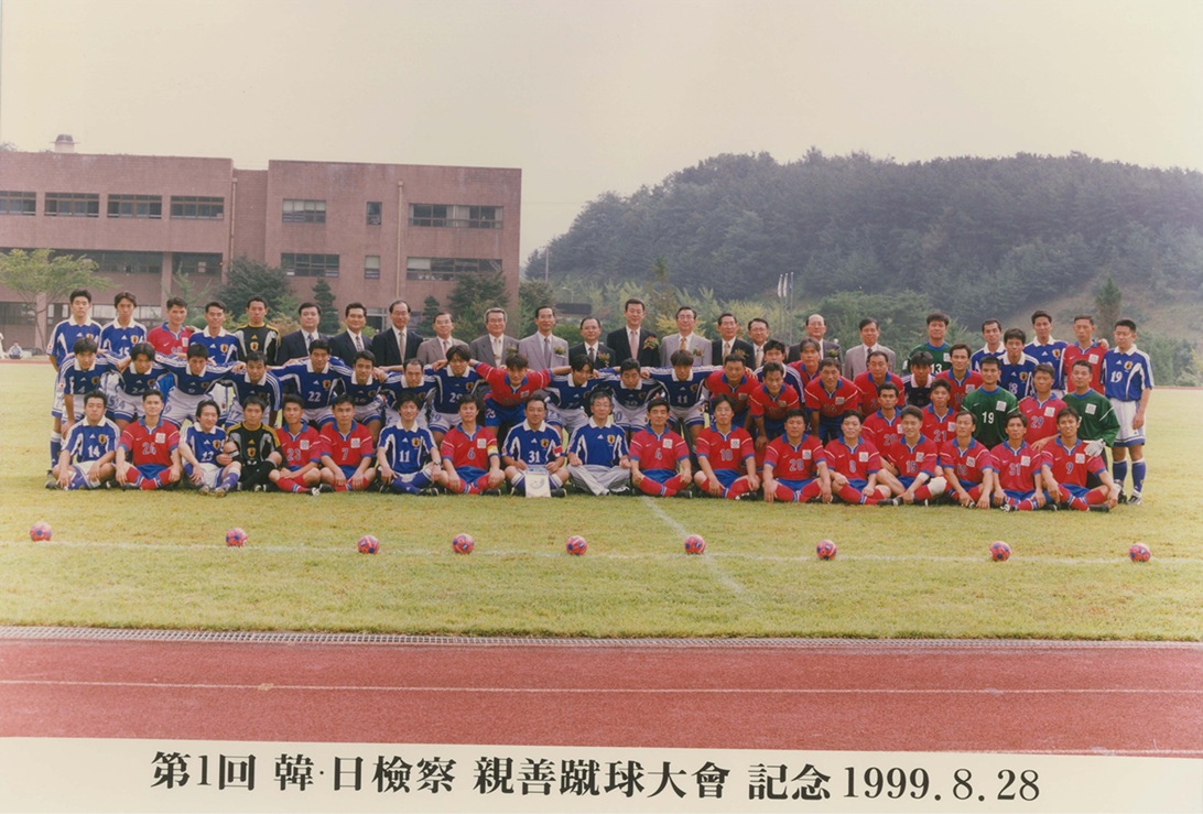 サッカーグラウンドを背景に、日本と韓国のユニフォーム姿の総勢70名ほどの男性が3列に並んでいる記念写真