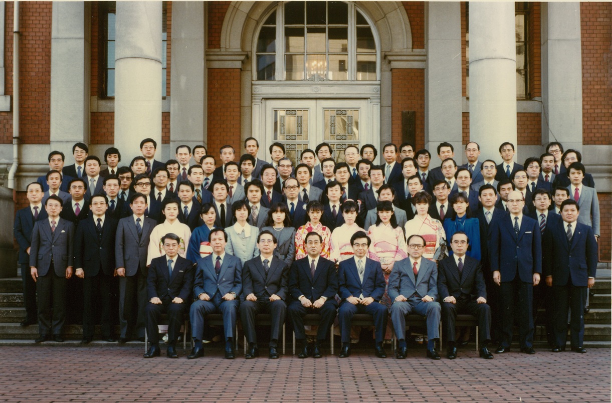 建物の前で、スーツ姿の男性と晴れ着の女性が並んでいる。ほとんどが男性で、女性の姿は少ない